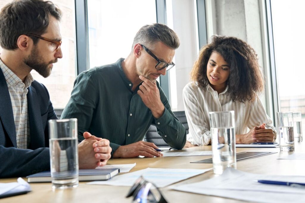 two businessmen and a businesswoman sitting at a desk in an office and reviewing a document