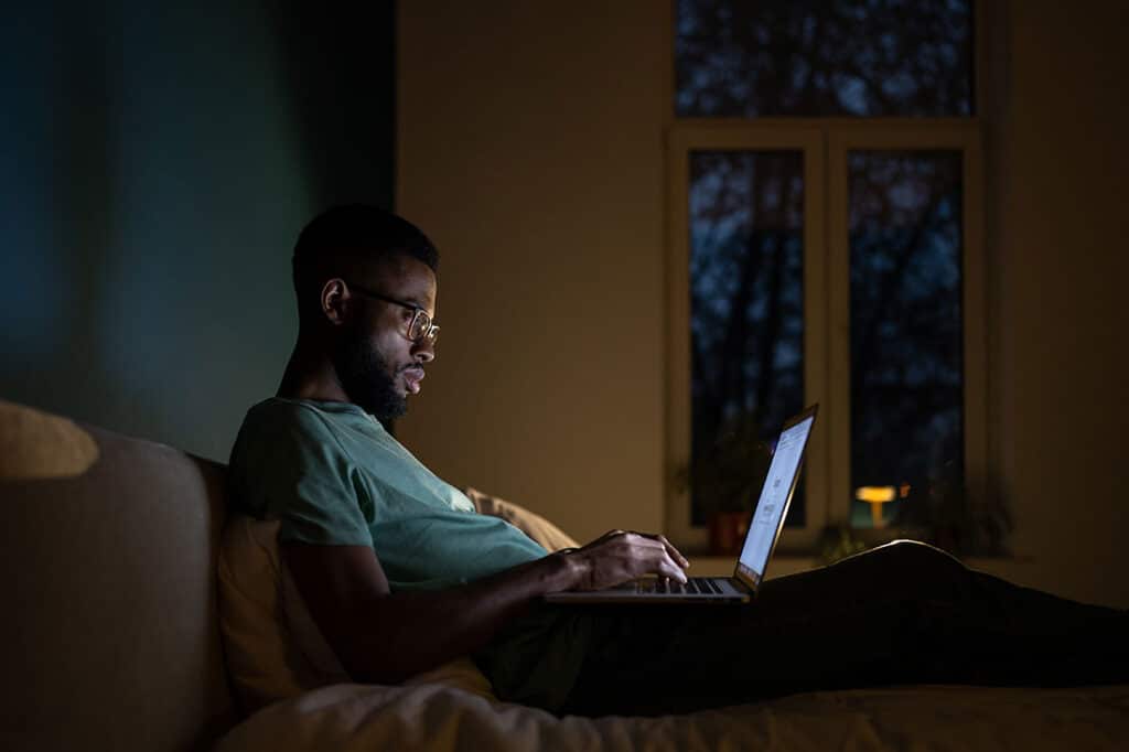 Focused man in dark room lying on bed work on laptop