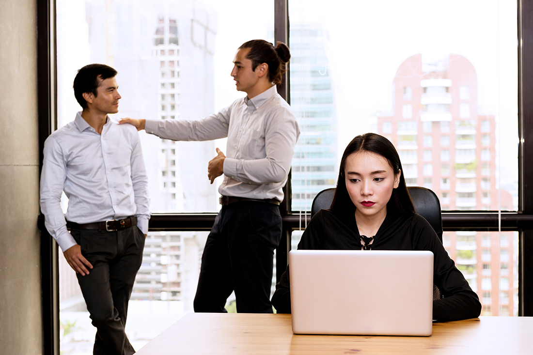Woman uses her laptop as two male coworkers communicate behind her in an office