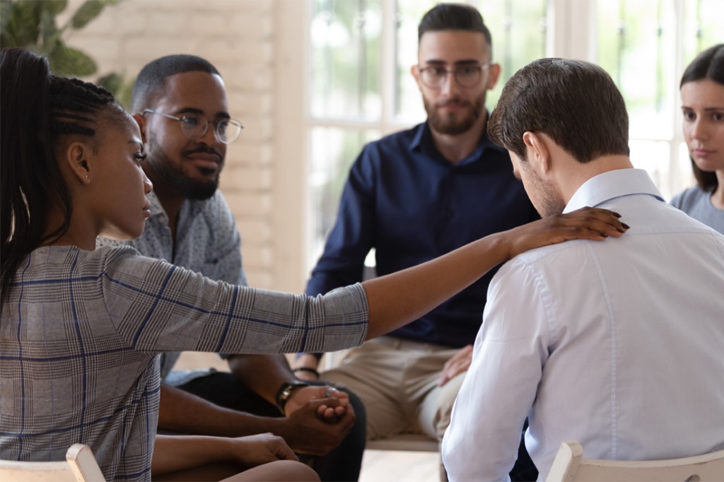 A support group, all in professional dress. A woman puts her hand on a man's shoulder to comfort him.